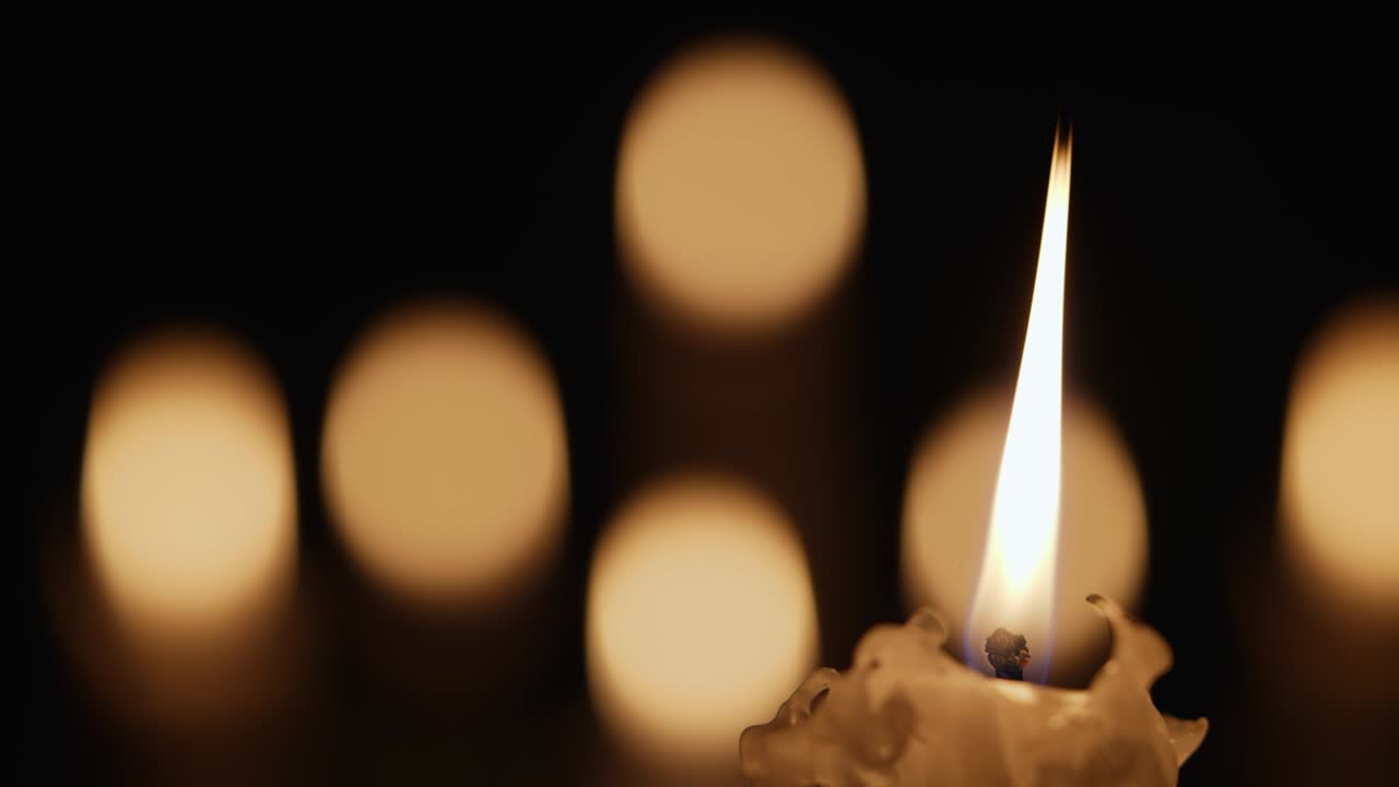 Candela di cera accesa che tremola nel buio su uno sfondo nero con effetto bokeh sfocato della luce della candela. Sfondo o illustrazione di ricordo, lutto o celebrazione. Primo piano di candele accese in chiesa