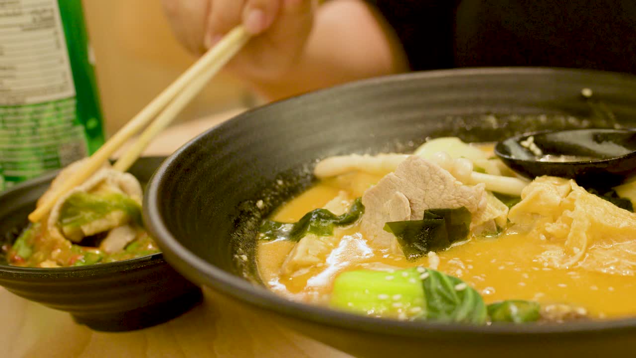 Una mano usa le bacchette per sollevare le verdure da una zuppa di noodle piccante in un ristorante caldo e con illuminazione soffusa