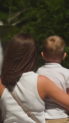 madre de cabello marrón y hijo pequeño descansan mirando una cascada en el parque de verano. joven y niño pasan tiempo cerca del río de montaña con cascada