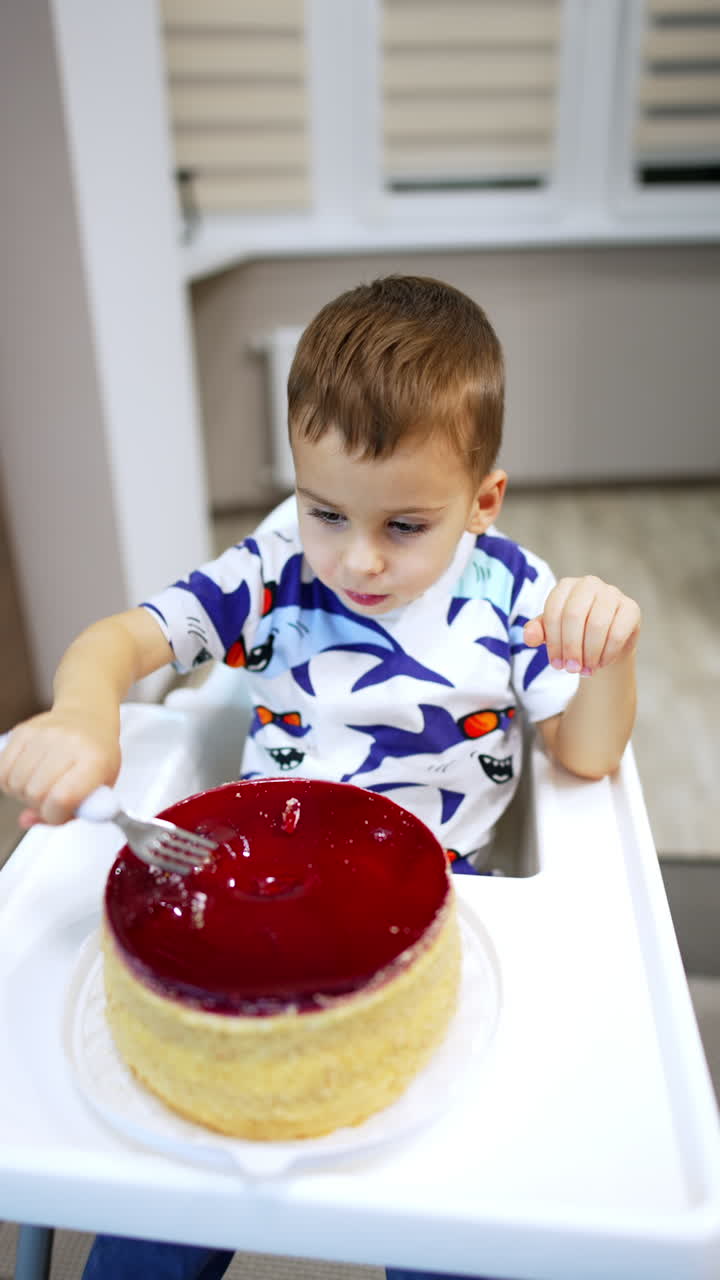 Lovely kid sitting at high chair scoops the cake. High angle view at the toddler eating a dessert. Vertical video.