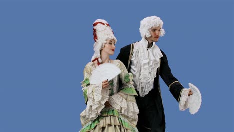 portrait of courtier lady and gentleman in historical vintage costumes and wig is waving a fan. young woman and man posing in studio with blue screen background. slow motion