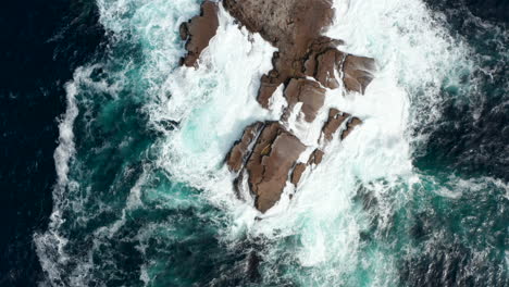 Aerial-birds-eye-overhead-top-down-panning-view-of-waves-crashing-on-coastal-rocks-and-making-white-foam.-Rough-sea-at-shore.-Kilkee-Cliff-Walk,-Ireland