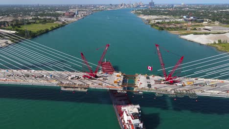 cargo vessel passing under newly built gordie howe international bridge with crawler cranes on construction site