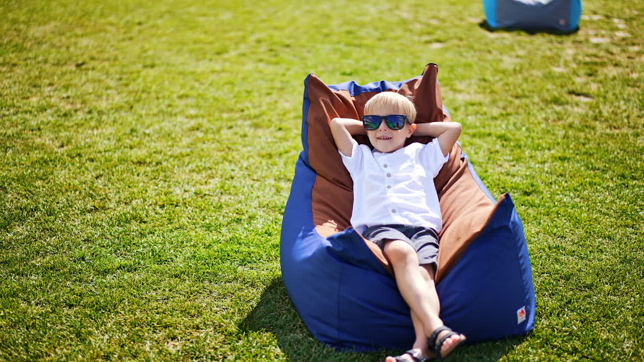 Little five-year old boy lies in bean bag chair outdoors in summer. Blond kid wearing sunglasses relaxing in the sun smiling happily.