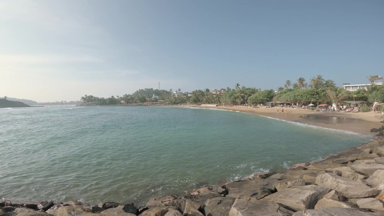 Massi in primo piano e una spiaggia orlata di palme scorrono attraverso l'inquadratura mentre una fluida panoramica a sinistra rivela le acque turchesi dell'Oceano Indiano e un piccolo isolotto al largo a Mirissa, Sri Lanka.
