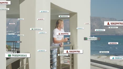 Animation-Von-Social-Media-Symbolen-Auf-Bannern-über-Einem-Kaukasischen-Mann,-Der-Auf-Einem-Balkon-Am-Meer-Kaffee-Trinkt