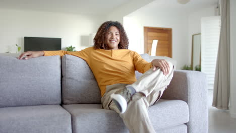 portrait of happy biracial man with long hair sitting on sofa smiling in living room, slow motion