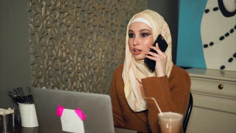young woman in hijab using laptop and phone in a cafe