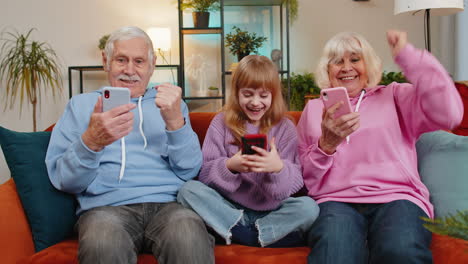 Granddaughter-and-grandparents-celebrating-victory-win-online-game-lottery-on-smartphones-at-home