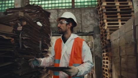 un hombre moreno con barba en un uniforme de protección blanco en un chaleco naranja camina a lo largo de las pilas con papel de basura y hace recuento en una gran planta de clasificación y reciclaje de residuos