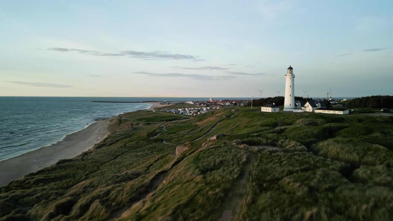 Fast evening drone flight over the dunes on the North Sea coast in Denmark at sunset. A white lighthouse becomes visible behind the grassy hills