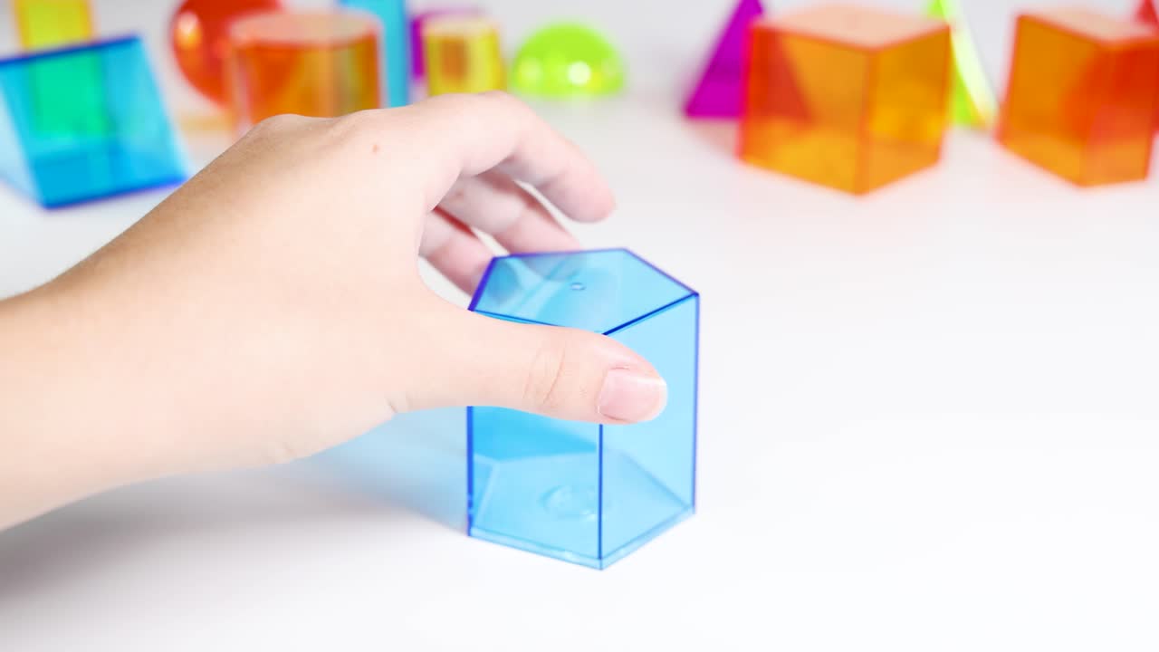 A hand manipulates a blue transparent cuboid among various colorful geometric shapes on a white surface