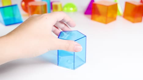 a hand manipulates a blue transparent cuboid among various colorful geometric shapes on a white surface