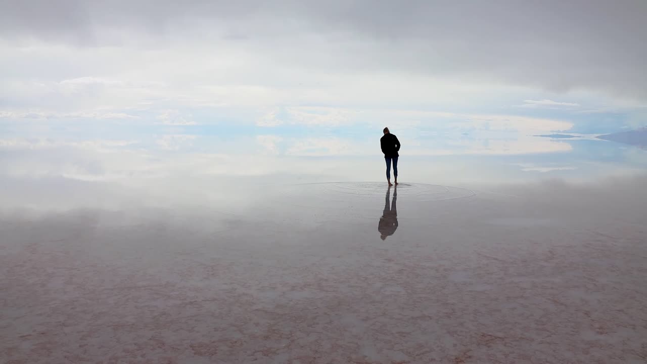 Lone silhouetted female walks on etheral Uyuni salt flats with mirror effect