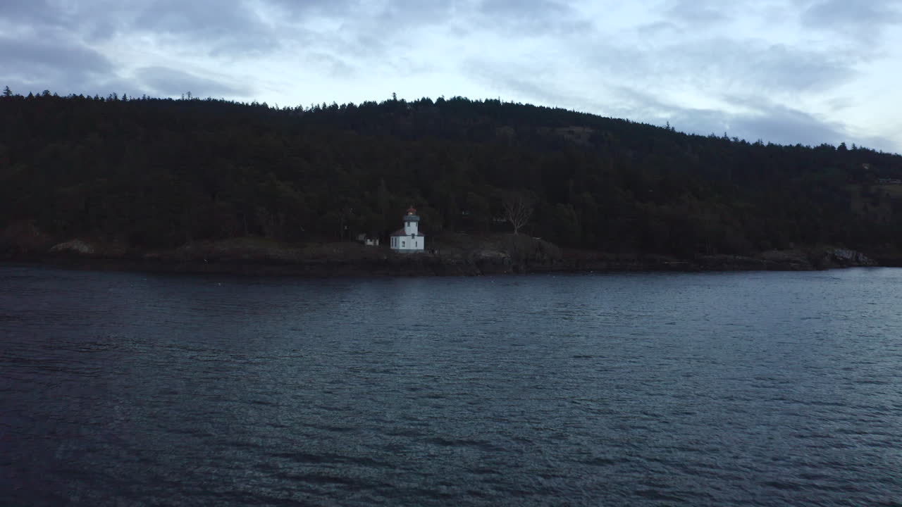Moody lighthouse drone. Drone flying toward lighthouse (wide shot) over water at sunset in the Pacific North West San Juan Islands. Birds flying over ocean with forest in background. Lighthouse 4k