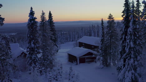 Aerial-view-moving-through-forested-alpine-trees-towards-remote-snow-covered-woodland-cabin-overlooking-wintertime-sunrise-landscape