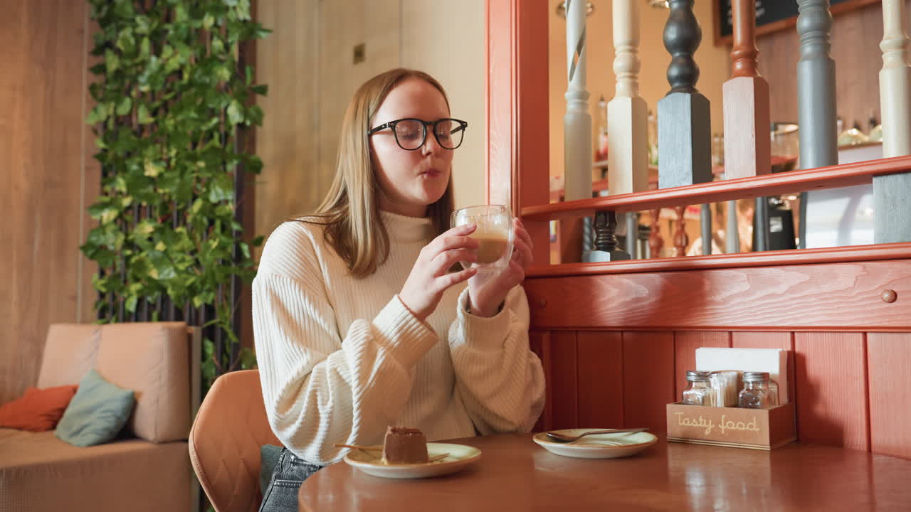 college student in white sweater sitting alone in cozy cafe lifts glass of latte while chocolate dessert sits nearby on plate, surrounded by warm lighting, red booth, and green indoor plants