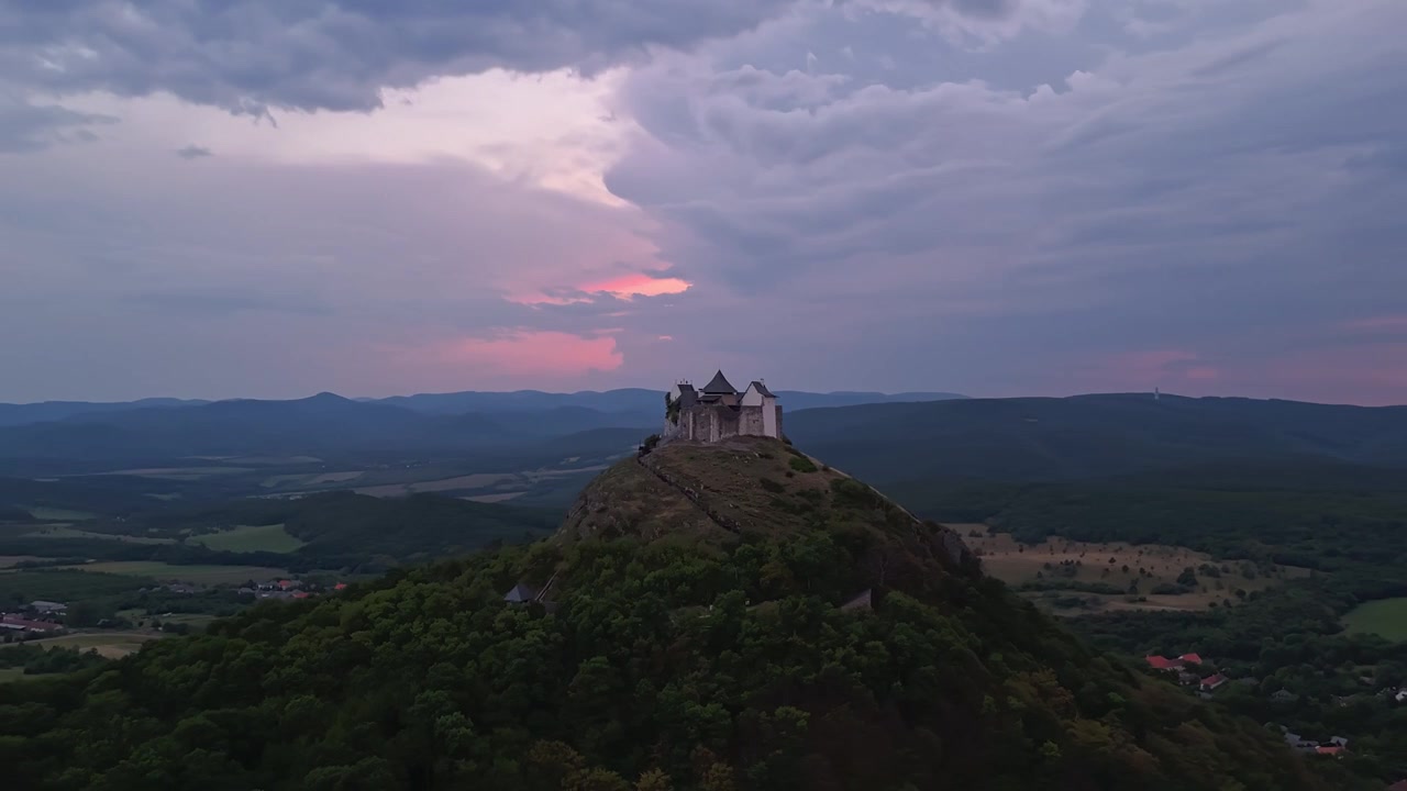 Füzér Slot i det nordlige Ungarn på en bakketop over de skovklædte Zemplén-bjerge under stormskyer og lyn ved skumringstid, med panoramaudsigt over landskabet i et drone-hyperlapse-perspektiv.