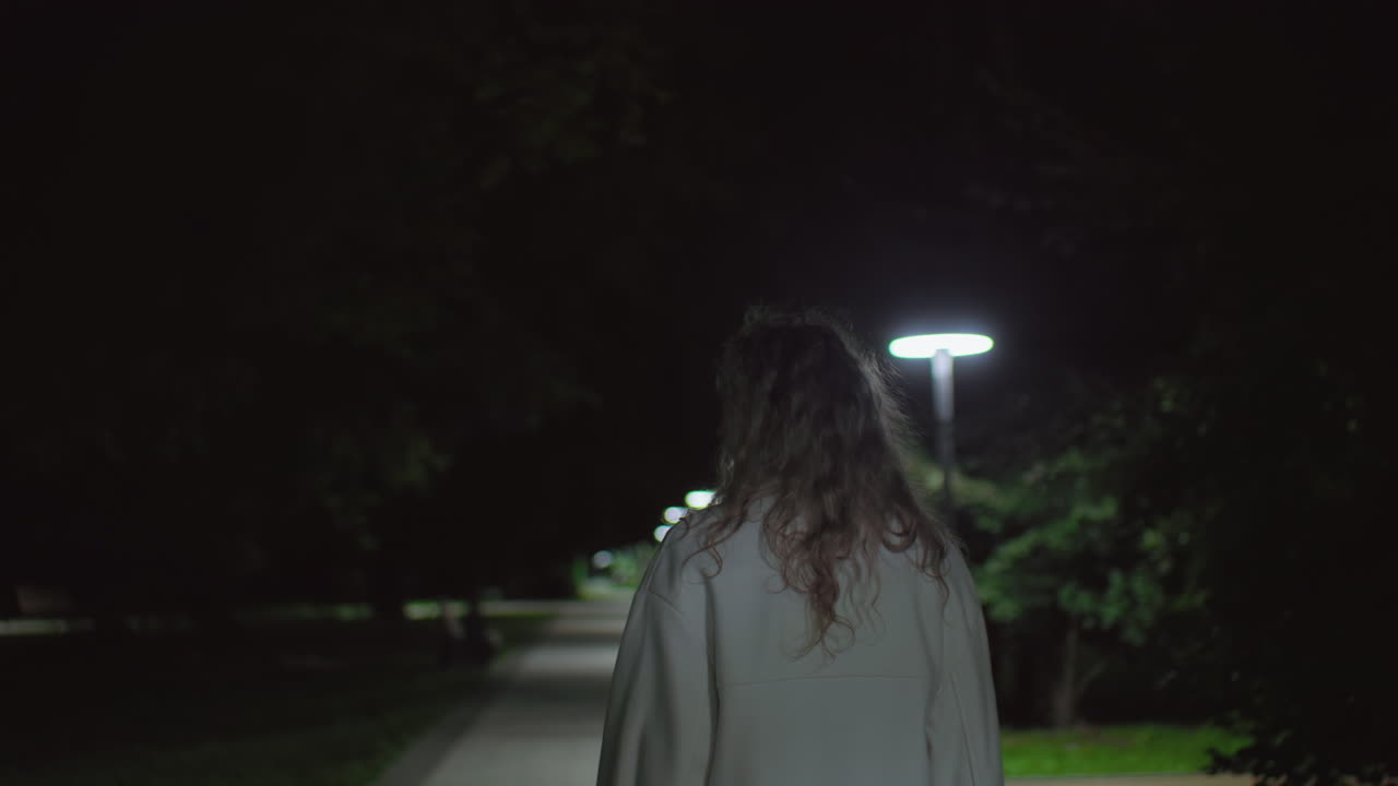 Back view young girl in white coat walking alone quietly at night along calm park walkway softly illuminated by gentle lights, creating atmosphere of peaceful solitude and quiet reflection