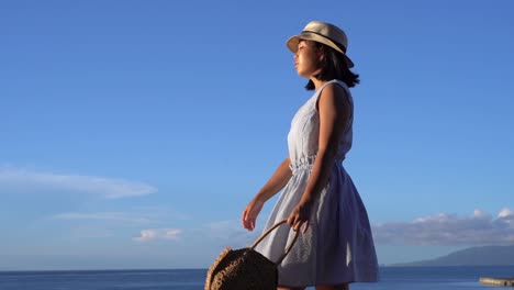 Chica-Japonesa-Con-Vestido-De-Verano-Sosteniendo-Una-Bolsa-Caminando-Frente-Al-Océano-En-Un-Hermoso-Día-Claro---Tiro-De-Seguimiento-Lateral-A-Cámara-Lenta