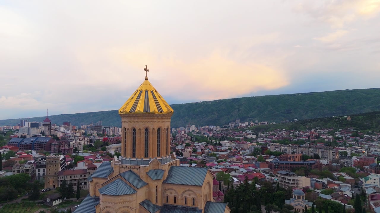 Detailní záběr dronem zlaté kupole a horní architektury Katedrály Nejsvětější Trojice (Sameba) v Tbilisi, Gruzie, s panoramatickým výhledem na město a okolní hory v pozadí.