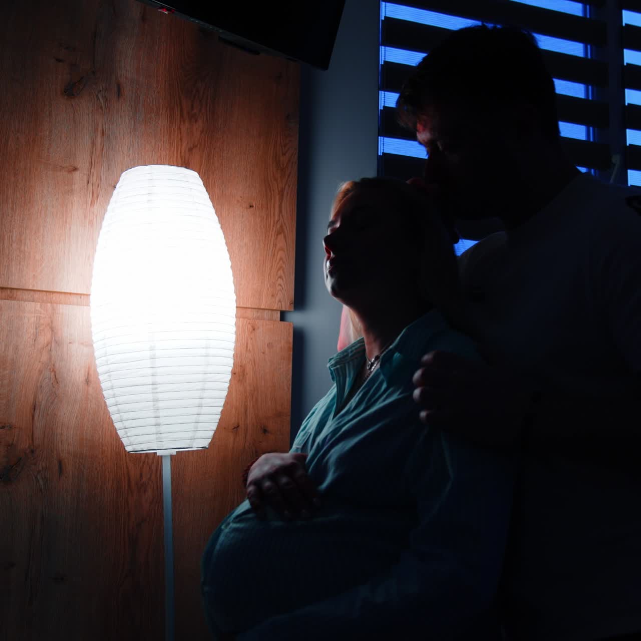 Loving husband holds and kisses his pregnant wife sitting on the bed. Woman strokes her big belly. Dark room with nightlight at backdrop