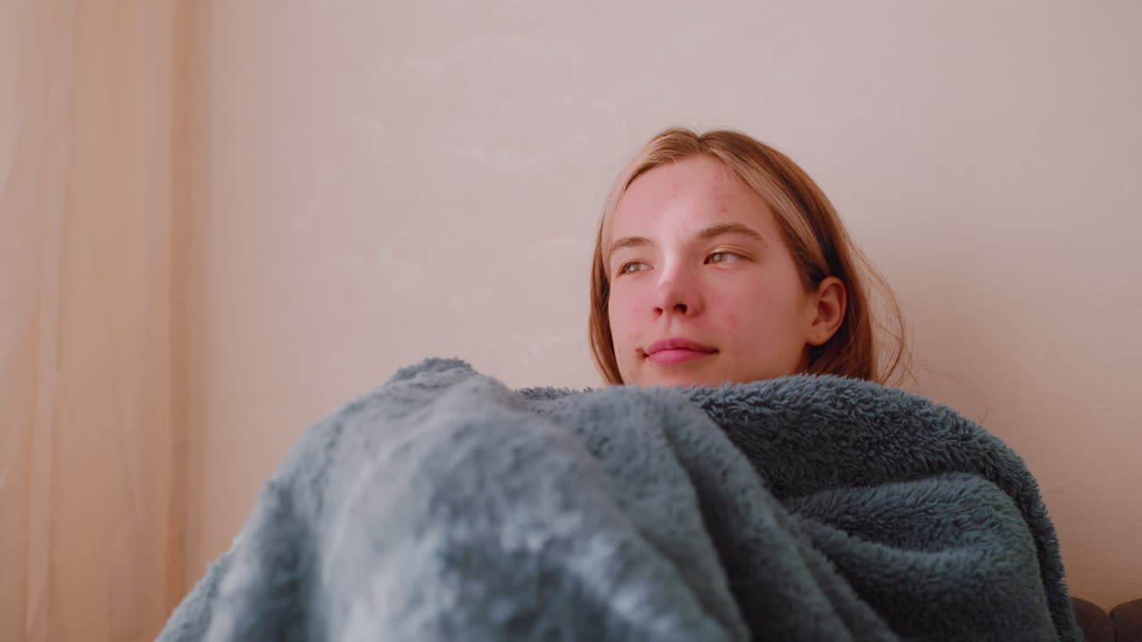 Independent woman seated on sofa covering herself with blanket while facing bright window with eyes gently closed enjoying calm atmosphere of warmth