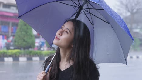 a young beautiful asian woman stands on roadside with holding a blue umbrella on rainy day caused by a tropical typhoon, waiting for bus on city roadside, weather forecasting, cold and flu prevention