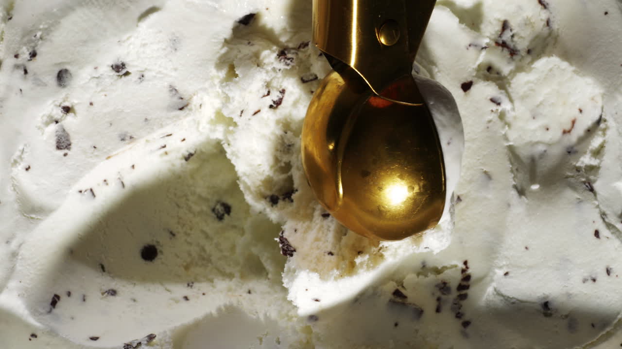 A shiny golden scoop glides into a tub of creamy chocolate chip stracciatella ice cream, capturing the rich texture and crunchy bits in a close-up view under soft lighting.