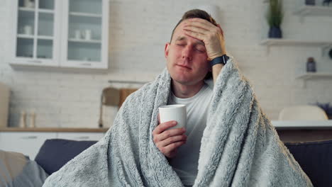 unhappy guy suffering coronavirus infection. sick man covered with warm blanket with a cup of hot beverage staying home. self-isolation and quarantine.
