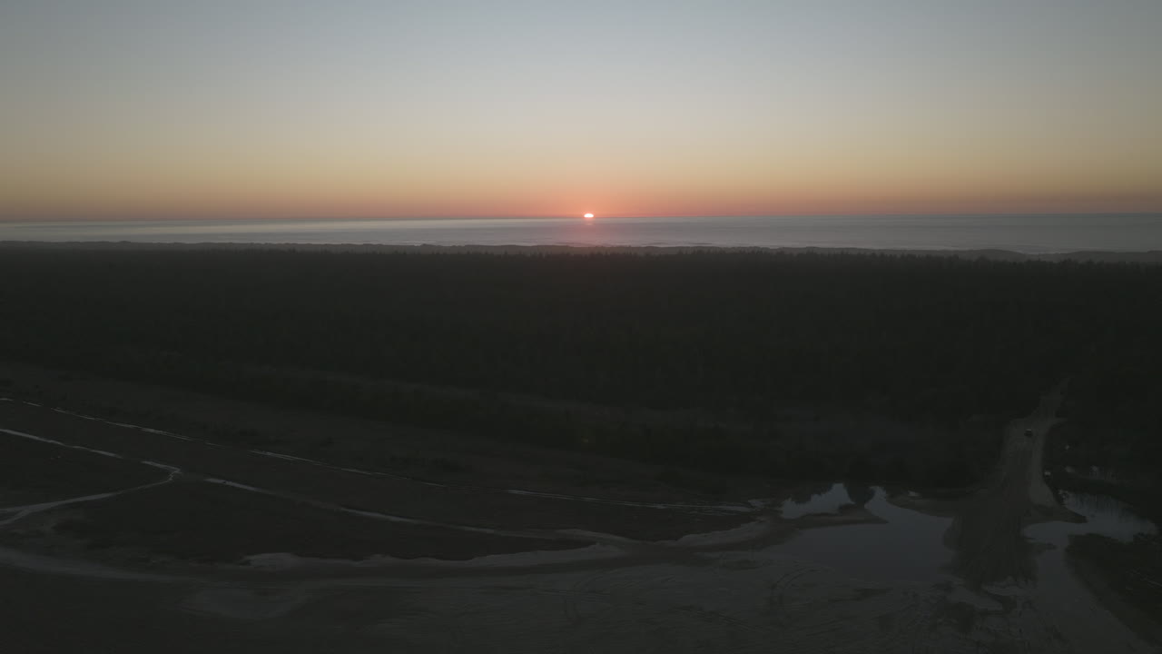Sun setting over sand dunes of Dunes City, Oregon, car drives along sand through trees towards ocean, aerial pullout.