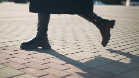 leg view of person in stylish leather boots and chic coat walking confidently on interlocked pavement under sunlight, shadow on ground, blurred background features parked cars