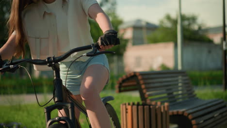 close-up of cyclist preparing for outdoor ride, mounting bicycle near colorful garden, adjusting hair under warm sunlight, surrounded by vibrant flower beds, trees, and wooden bench