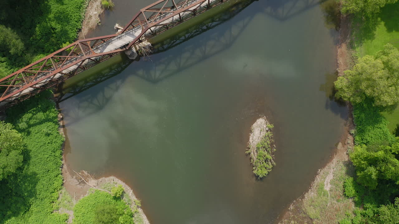 drone aéreo volando sobre el río azul reflectante y el viejo puente oxidado junto a un bosque verde brillante en el centro de pensilvania durante el verano