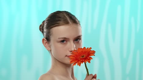 young woman holding gerbera flower with gentle smile a young woman with a gentle smile holds a vibrant orange gerbera flower against a serene blue background