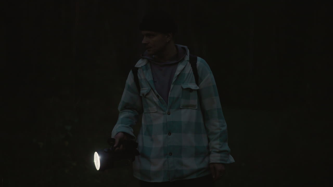 Excursionista nocturno en un bosque con linterna explorando un sendero oscuro, ambiente cinematográfico melancólico, figura solitaria con chaqueta impermeable y mochila, haz de luz atravesando la niebla, mirada ansiosa hacia arriba, aislamiento lleno de suspense