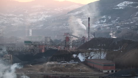 Der-Abend-Bricht-über-Einen-Industriekomplex-Inmitten-Einer-Berglandschaft-Herein,-Aus-Einem-Schornstein-Steigt-Sanft-Rauch-In-Den-Himmel