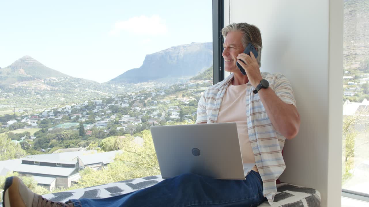 Getting call mature man holding phone, talking while working on laptop at window seat, copy space