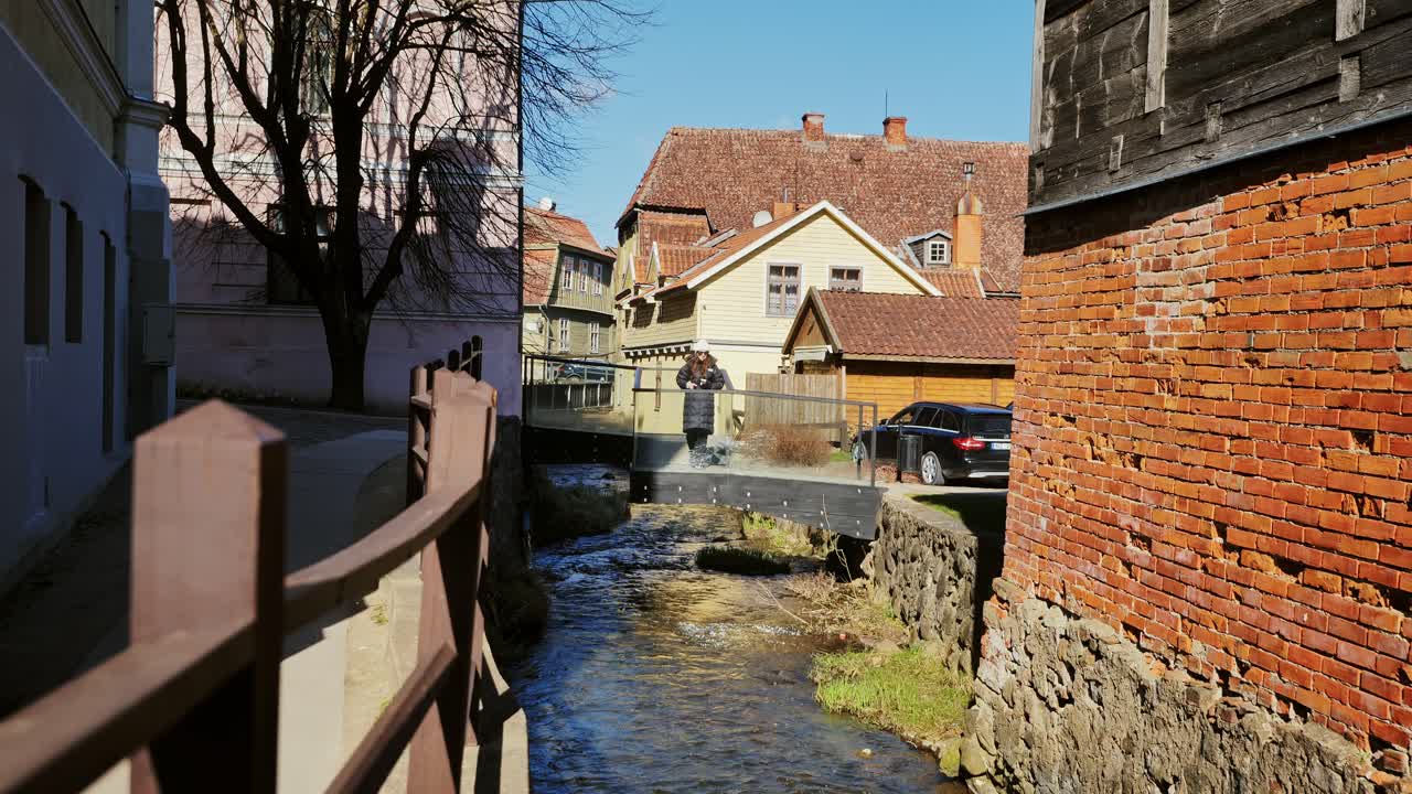 Woman stands calmly on old bridge filming gentle stream through Kuldīga town