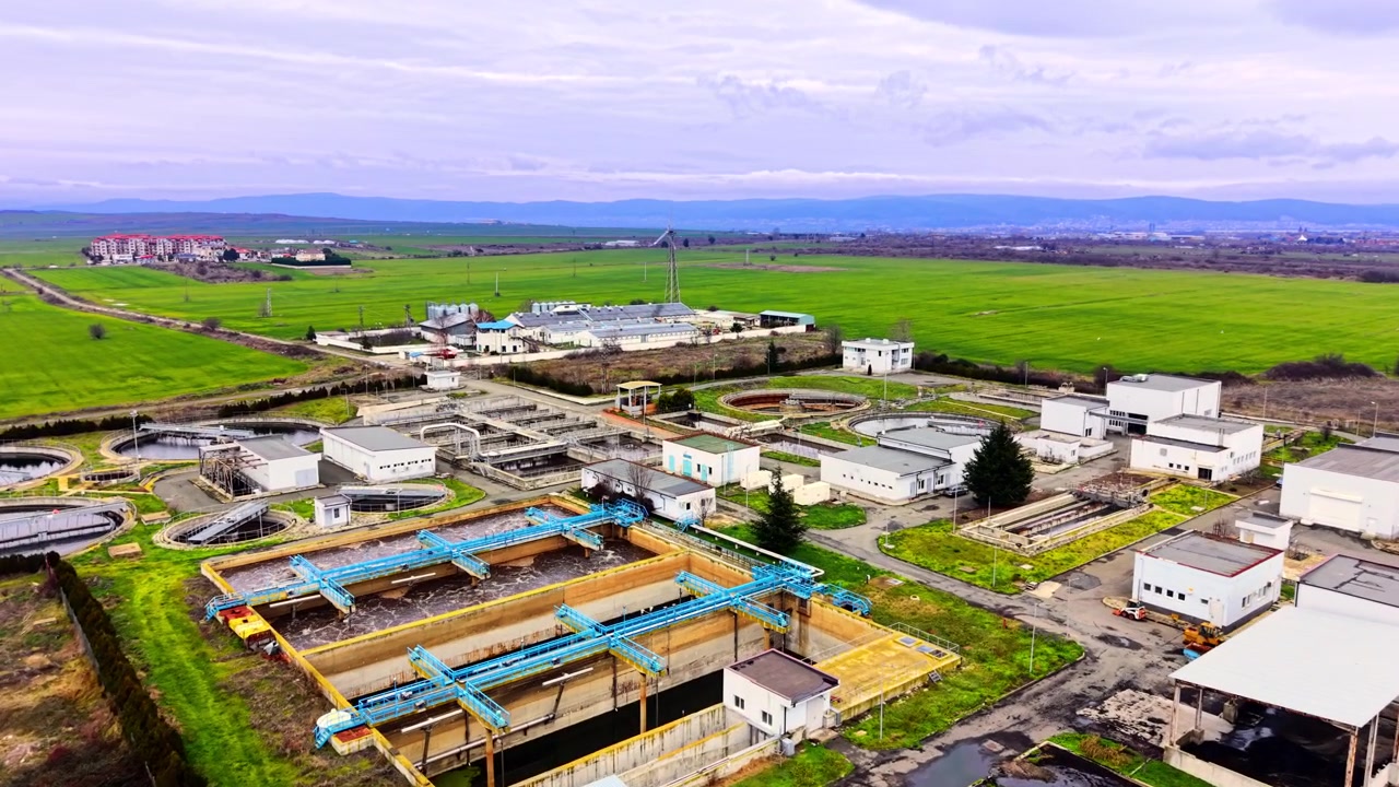 Water treatment plant sits amid green farmland. Aerial view of circular tanks, blue pipework and white buildings surrounded by open fields.