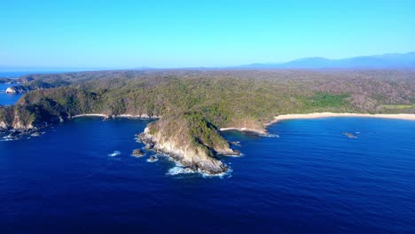 Strafe-Flight-along-Rocky-Coastline-of-Dark-Blue-Ocean,-Large-Open-Beach-and-Distant-Mountains