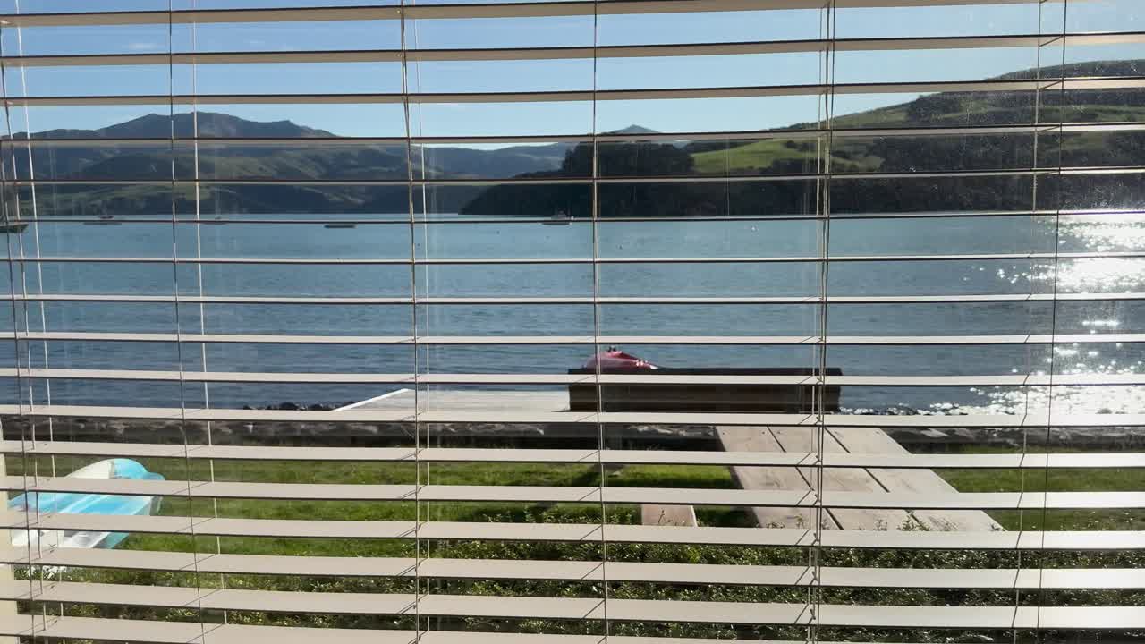 A tranquil lakeside scene viewed through window blinds, capturing sunlight reflecting on water in Akaroa, New Zealand