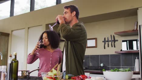 happy diverse couple preparing a meal together in kitchen, drinking wine and embracing