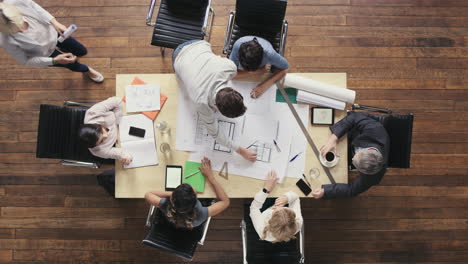 Vista-Superior-De-Gente-De-Negocios-Reunida-Alrededor-De-Una-Mesa-De-Juntas-Discutiendo-Planes-Arquitectónicos-Para-Un-Nuevo-Espacio-De-Oficinas-Compartido-Sostenible