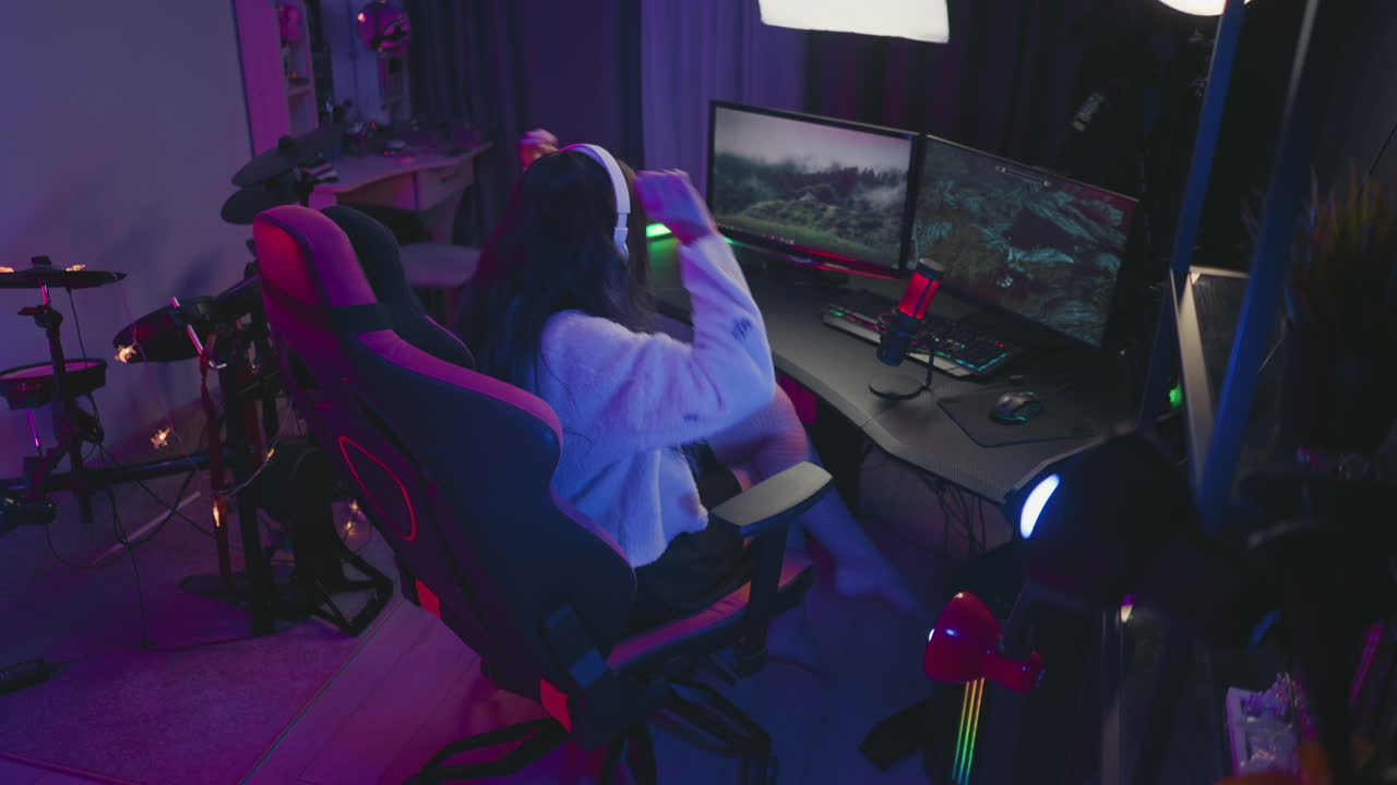 back view of focused female gamer seated in illuminated room using dual monitors and gaming gear, surrounded by glowing lights, drum set, microphone, and streaming setup