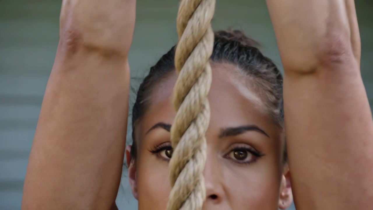Gripping thick rope, female athlete pulling upward at building siding for training in camo tank