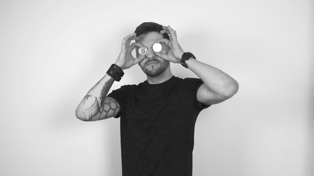 Grayscale footage of a tattooed short haired bearded man lifting up two bitcoin coins with his hands and places them in front of his eyes in a funny way. Coins are reflective in the light and pretty.