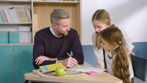 profesor sentado en el escritorio resolviendo dudas a dos alumnas en el aula de inglés 1