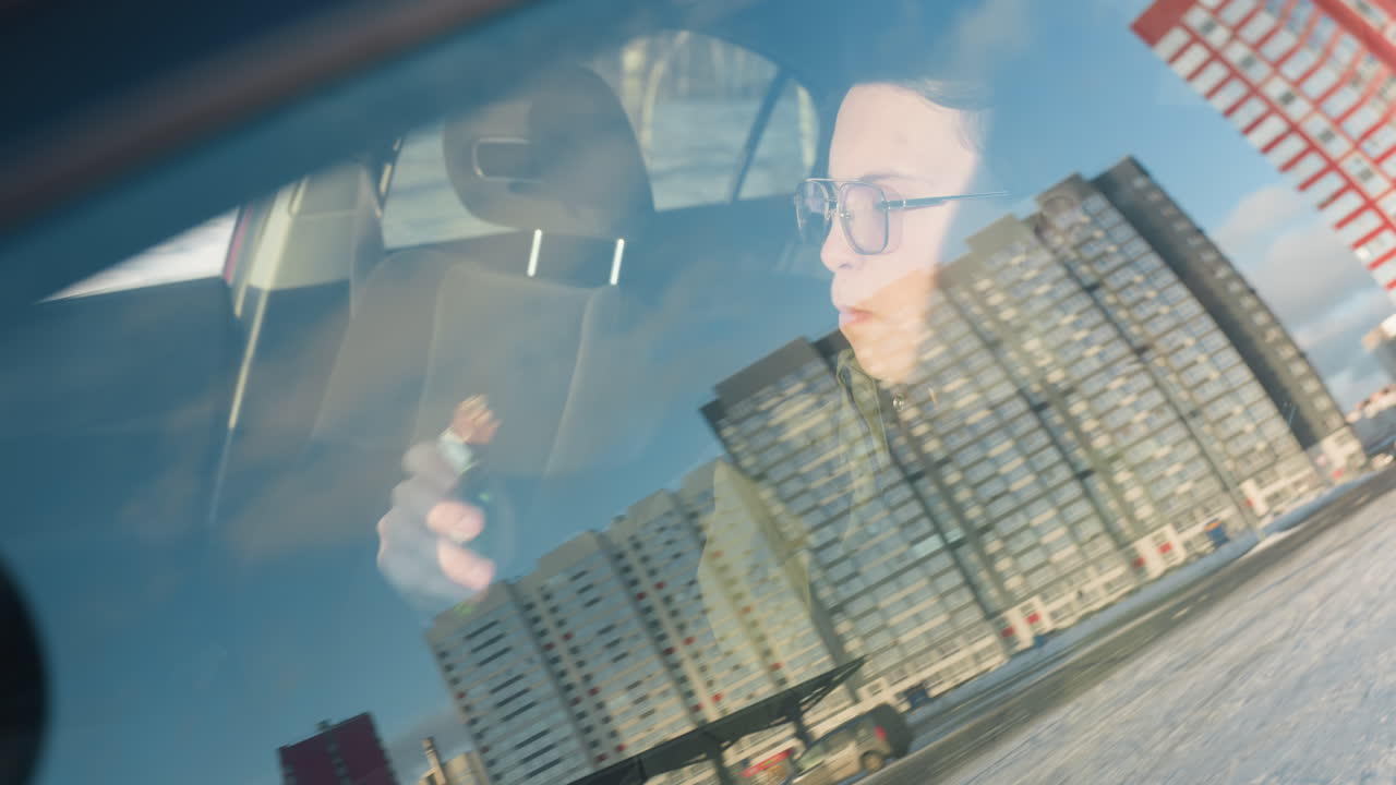 close up through car window reflecting office building view as student eats chocolate and sips juice inside parked vehicle with blurred urban winter backdrop under soft daylight