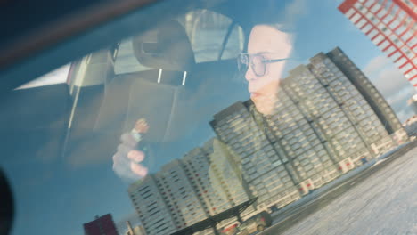 close up through car window reflecting office building view as student eats chocolate and sips juice inside parked vehicle with blurred urban winter backdrop under soft daylight